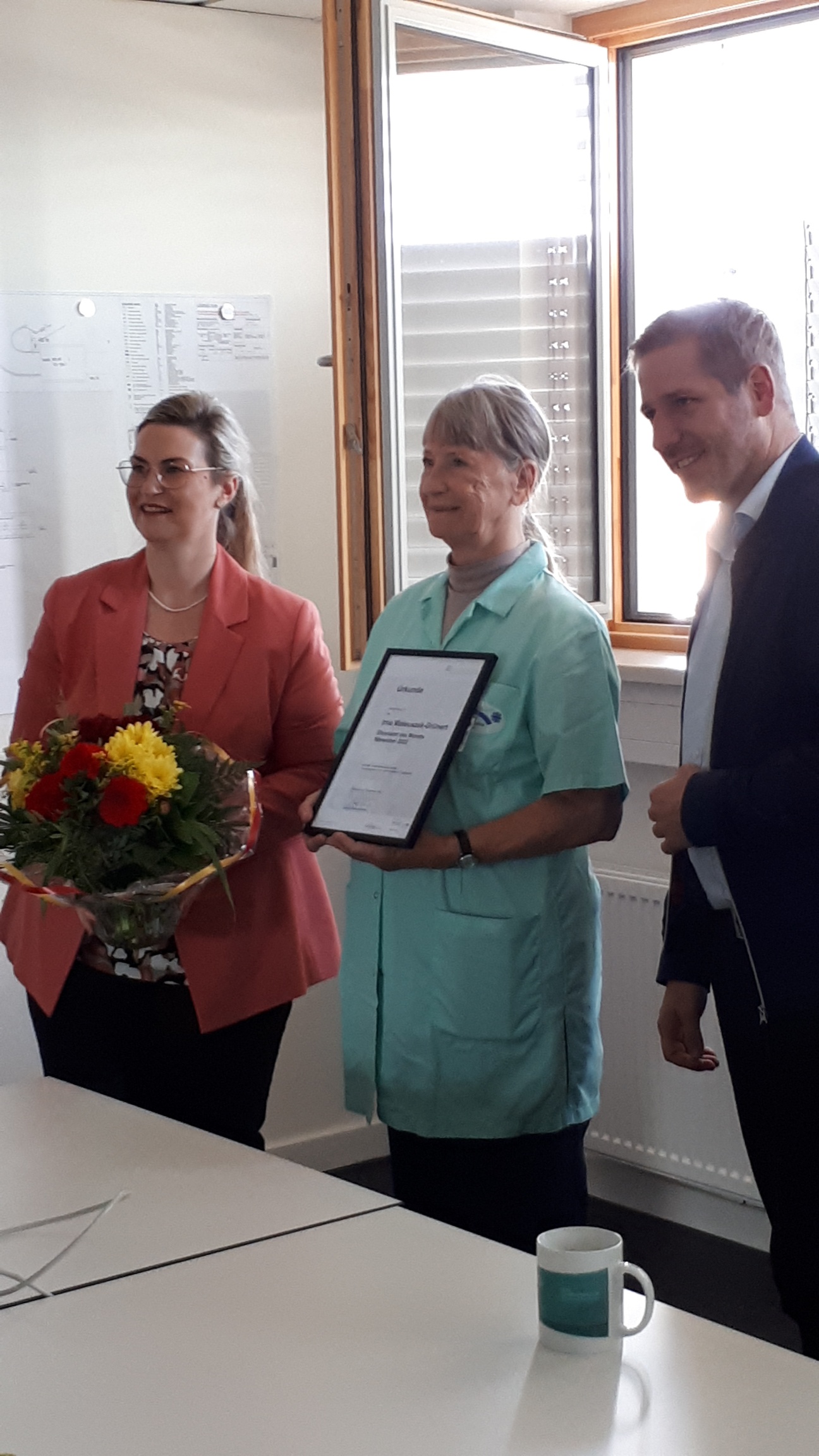 Zwei Personen stehen neben einer Dame, die eine Urkunde hält